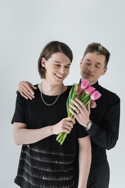 Молодой гей улыбается, держа цветы рядом с парнем, изолированным от серых  - Фото, изображение