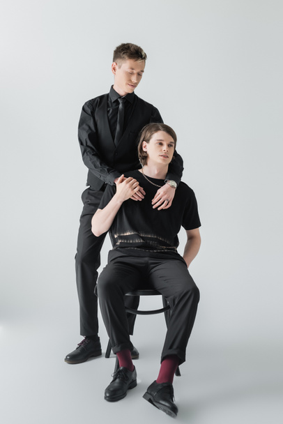 Stylish gay embracing boyfriend sitting on chair on grey background  - 写真・画像
