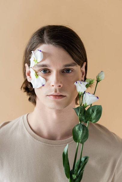 Brunette homme avec fleur sur le visage tenant eustome isolé sur beige  - Photo, image