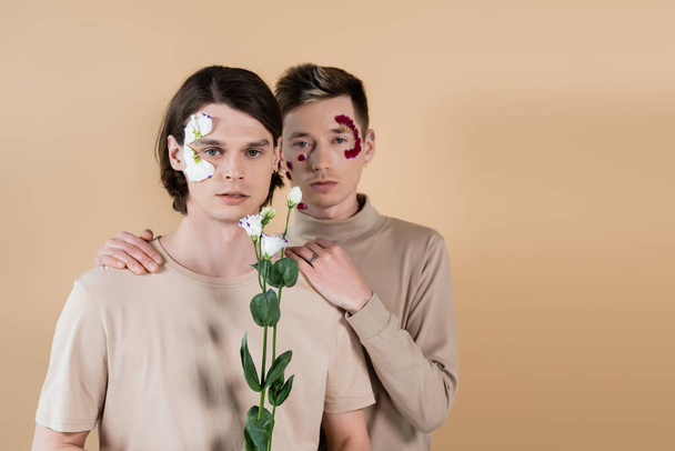 Gay with petals on face hugging boyfriend with eustoma isolated on beige  - Photo, Image