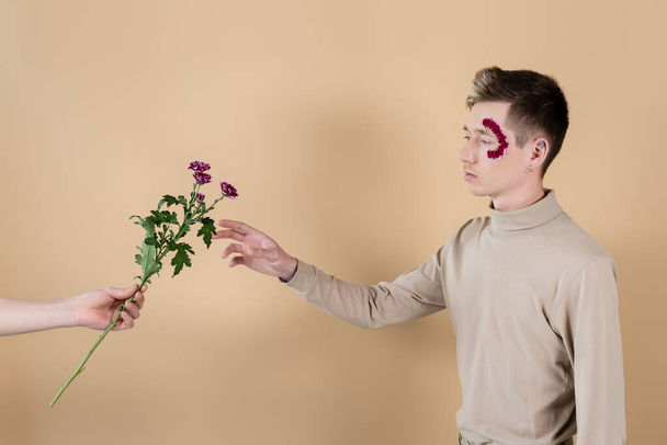 Mann hält Chrysanthemen neben Freund mit Blütenblättern im Gesicht auf beige  - Foto, Bild