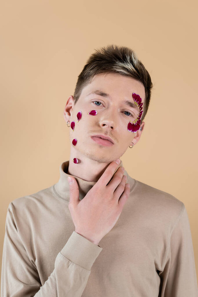 Портрет молодого человека с лепестками на лице, касающимися шеи, изолированный на бежевом  - Фото, изображение