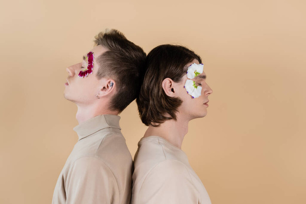 Side view of gay couple with petals on faces standing back to back isolated on beige  - 写真・画像