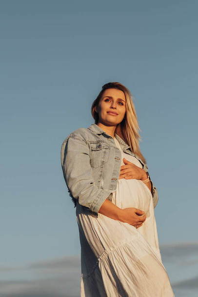 Retrato de una joven mujer caucásica embarazada sosteniendo su vientre sobre el fondo del cielo azul - Foto, Imagen
