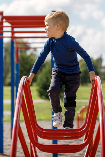Un bambino si arrampica e pende su una scala metallica a forma di arco nel parco giochi. - Foto, immagini