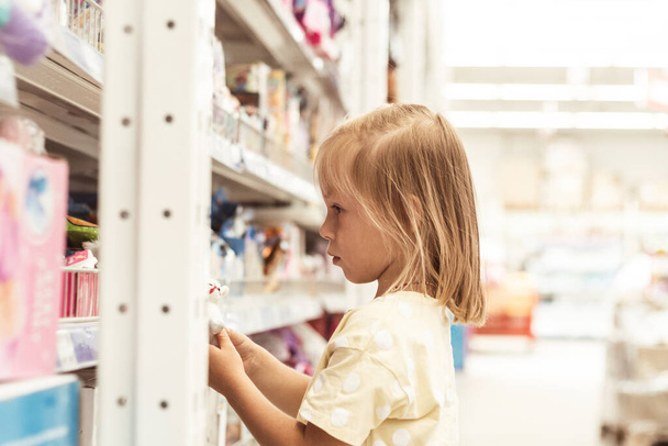 Little caucasian girl choosing a new toy in the big baby store. Big shelfs full of toys - Foto, Bild