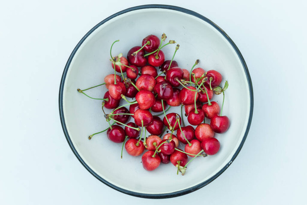 Many red cherry fruits in round bowl, isolated on white. Organic berries fresh. A lot of sweet cherries with petioles top view - Foto, afbeelding