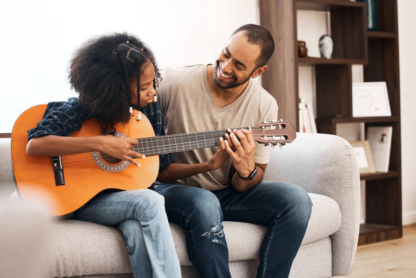Shot van een jong meisje leren gitaar spelen met haar vader thuis. - Foto, afbeelding