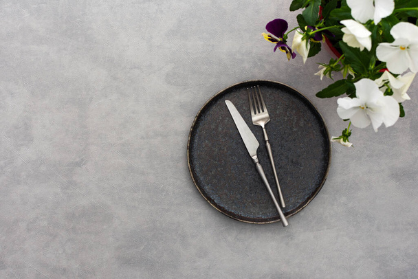 Tischdekoration, leerer Teller und Besteck auf grauem Hintergrund, Ansicht des Serviertisches mit Stiefmütterchenblumen - Foto, Bild