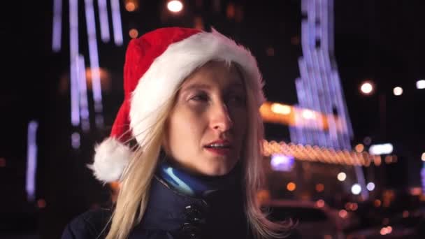 Een jonge vrouw met een kerstmuts staat met haar mond wijd open. Achter het is een nachtstad met iriserende lichten van winkelpuien, advertenties en bloemenslingers. Ze is opgewonden en overstuur.. - Video