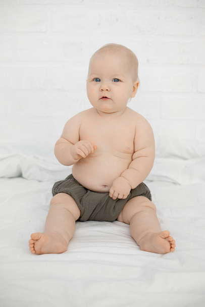 Pretty little baby sit on big bed with satisfied facial expression on white background. Portrait of beautiful naked infant child, free copy space. Concept of happy childhood and caring for children. - Φωτογραφία, εικόνα