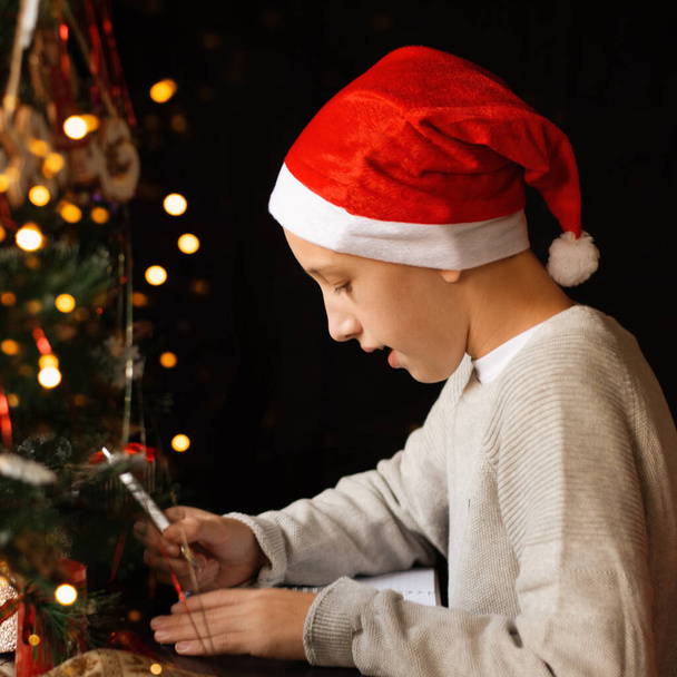 The boy in the Santa hat sitting near the Christmas tree and write a wish. Blurred lights. Close up. Copy space. - Photo, Image