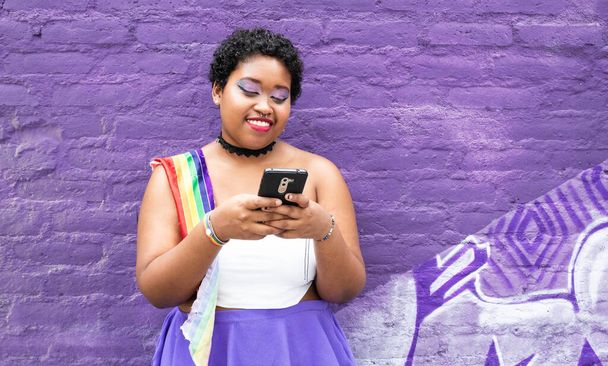 lgbt afrikanisch-amerikanische Mädchen mit Handy im Chat mit lgbt Flagge in der Stadt auf lila Wand Hintergrund lächelnd - Foto, Bild