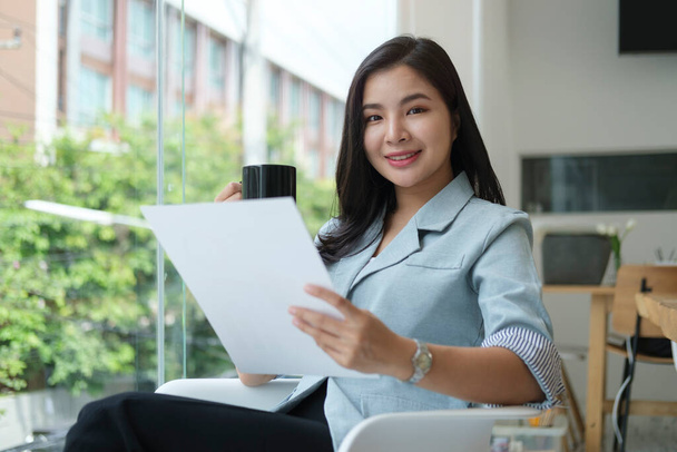 Smiling asian businesswoman sitting in bright modern office and reading document. - Foto, imagen