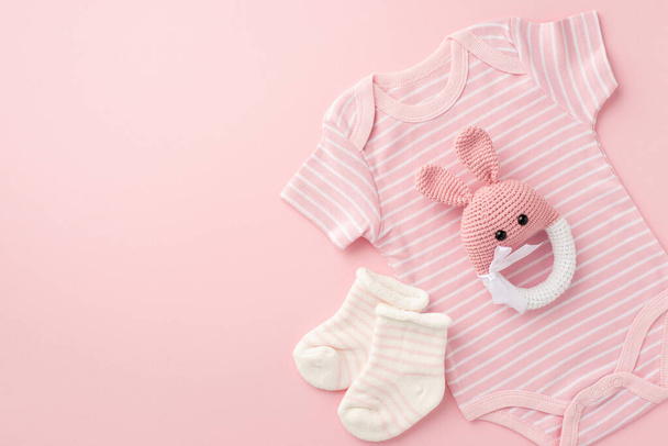 Concepto de bebé. Foto vista superior de ropa infantil calcetines de body rosa y juguete de sonajero de conejito de punto sobre fondo rosa pastel aislado con espacio vacío - Foto, Imagen