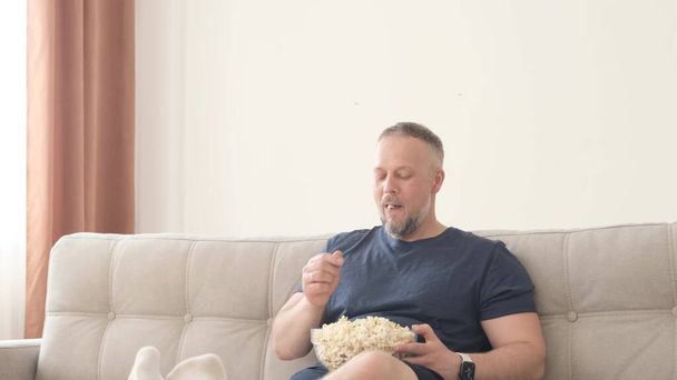 The man is relaxing at home on the couch, he is eating popcorn and watching TV. Happy relaxed guy watching movie program funny TV series online eating popcorn laughing having fun at home - Photo, Image