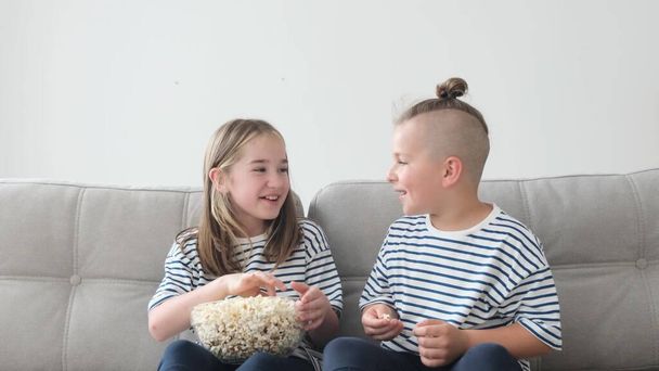 Брат и сестра едят попкорн, сидя на диване, выбрасывают его и ловят ртом. Двое детей смотрят телевизор и едят вкусный попкорн - Фото, изображение