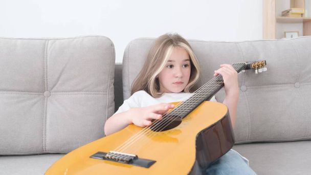 Triste niña tocando la guitarra, ella está descansando sentada en el sofá en casa. Chica músico adolescente. Vídeo 4k - Foto, imagen