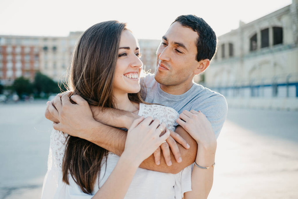 Een Spaanse man knuffelt zijn lachende brunette vriendin terwijl ze 's avonds in de haven van Spanje lopen. Een paar toeristen op een date bij zonsondergang in Valencia. - Foto, afbeelding