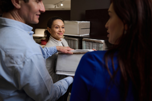 Close-up of a married couple in a furniture store inspects the orthopedic mattress sample before buying, standing near a pleasant experienced sales consultant. Healthy sleep - Photo, Image