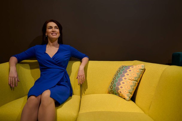 Superbe séduisante femme caucasienne ravie en robe bleue formelle testant un entraîneur d'art nouveau jaune dans un studio de design de meubles. Concept de design d'intérieur et de maison - Photo, image