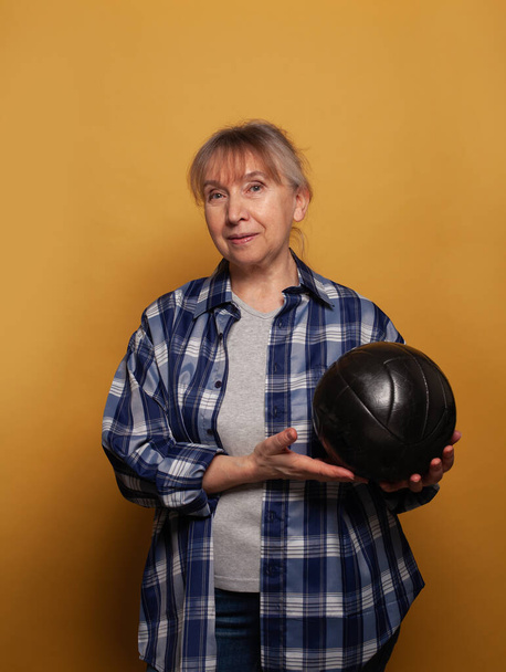 Smiling senior woman in casuals holding soccer ball against yellow background - Foto, afbeelding
