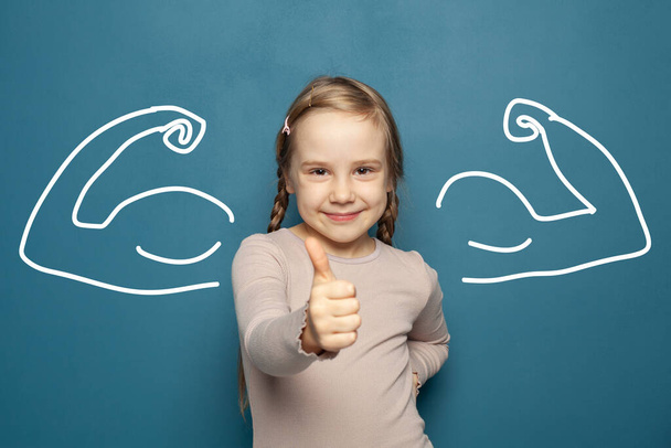 Nettes fröhliches Kind Mädchen zeigt Daumen nach oben gegen Muskeln auf blauem Hintergrund - Foto, Bild