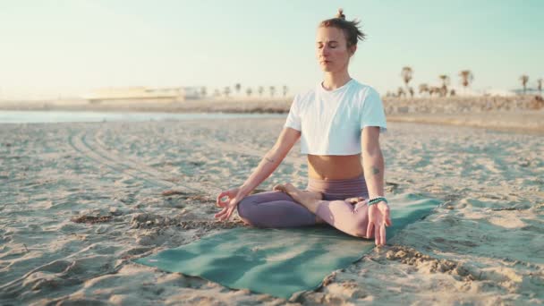 Linda menina loira yogi vestida de sportswear sentado em lótus posar no tapete junto ao mar. Jovem mulher tatuada meditando na praia. Yoga manhã ao ar livre - Filmagem, Vídeo