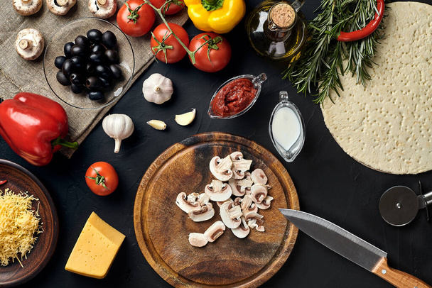 Cutting wooden board with traditional pizza preparation ingredients: cheese, tomatoes sauce, mushrooms, olive oil, pepper, spices. Black texture table background. Top view. Still life. Copy space - 写真・画像