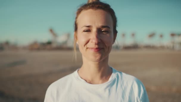 Κοντινό πλάνο όμορφη ξανθιά γυναίκα κοιτάζοντας κάμερα και χαμογελώντας ξοδεύοντας χρόνο στην παραλία. Νεαρή σπορ γυναίκα που ποζάρει σε εξωτερικούς χώρους - Πλάνα, βίντεο