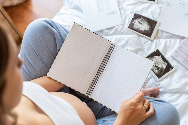 Контрольный список беременных женщин. Красивая беременная женщина выписывает чек-лист. Счастливая беременная женщина держит блокнот. Концепция материнства, ожидания - Фото, изображение