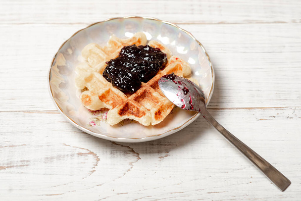 Belgian waffles with blueberry or blackcurrant jam on a plate with a spoon on a white wooden background. - Foto, Imagem