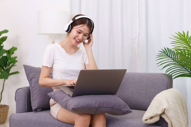 Életmód a nappaliban koncepció, fiatal ázsiai nő fejhallgató zenét hallgatni laptop. - Fotó, kép