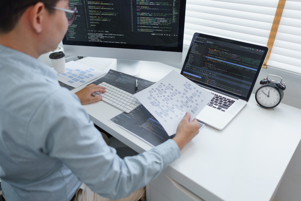 Η έννοια ανάπτυξης λογισμικού, αρρενωπός προγραμματιστής διαβάζει τα στοιχεία στο έγγραφο ενώ προγραμματίζει στον υπολογιστή. - Φωτογραφία, εικόνα