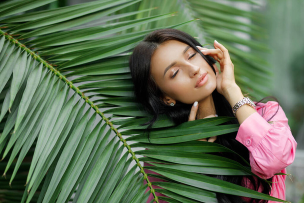 Portrét mladé ženy se zavřenýma očima, bruneta v růžových šatech pózující v tropické botanické zahradě s palmami na pozadí. - Fotografie, Obrázek