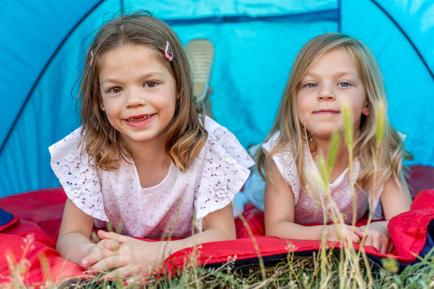 portrait of two blonde and pretty sisters lying down in a blue camping tent - summer vacation concept - Foto, Bild