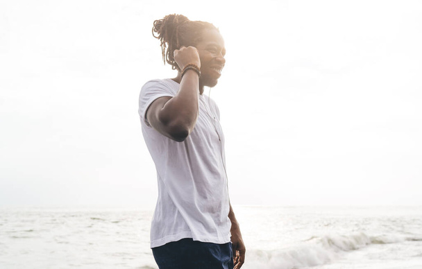 From below of content African American male in casual clothes smiling and looking away while chilling on seashore with music - Foto, imagen