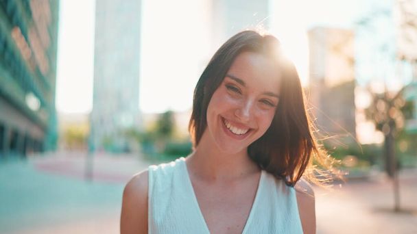 Cute young woman with freckles and dark loose hair wearing white top and jeans is smiling on modern city background. Beautiful girl with long eyelashes smiling in the back light - Photo, image