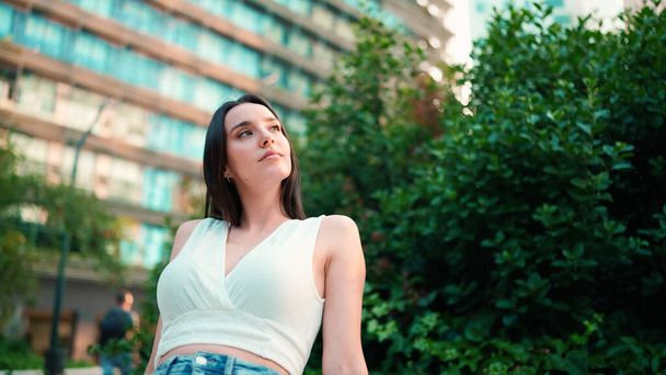Young woman with freckles and dark loose hair wearing white top and jeans is enjoying the view of modern city. Girl with long eyelashes sits in public garden on modern building background - Photo, image