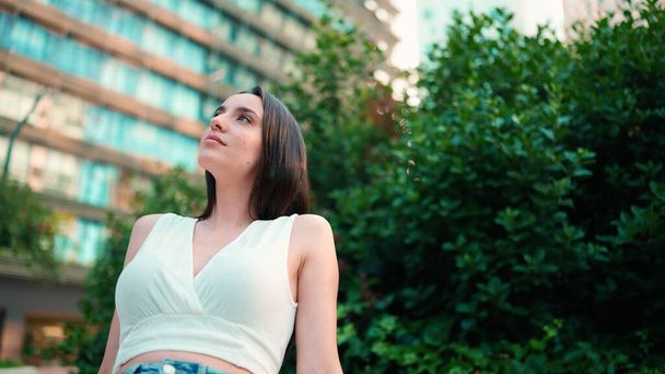 Young woman with freckles and dark loose hair wearing white top and jeans is enjoying the view of modern city. Girl with long eyelashes sits in public garden on modern building background - Photo, Image