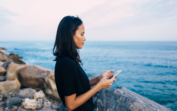 Vue latérale d'attrayant millénium féminin en utilisant un appareil smartphone pour vérifier l'emplacement du littoral pendant les vacances d'été - Photo, image