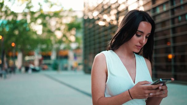 Beautiful woman with freckles and dark loose hair wearing white top is walking down the street with smartphone in her hands. Girl uses mobile phone on modern city background - Photo, image