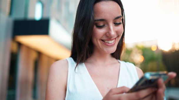 Beautiful woman with freckles and dark loose hair wearing white top is walking down the street with smartphone in her hands. Girl uses mobile phone on modern city background - 写真・画像