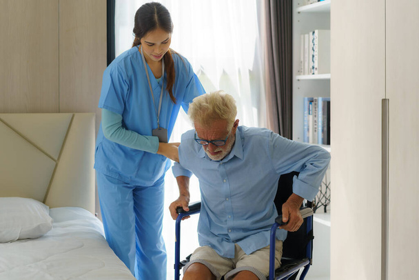 Azjatycka uśmiechnięta pielęgniarka pomaga starszemu mężczyźnie wstać z łóżka, lecząc pacjenta podczas wstawania z łóżka i poruszania się na wózku inwalidzkim w domu, pomagając niepełnosprawnym starszym osobom wstać - Zdjęcie, obraz