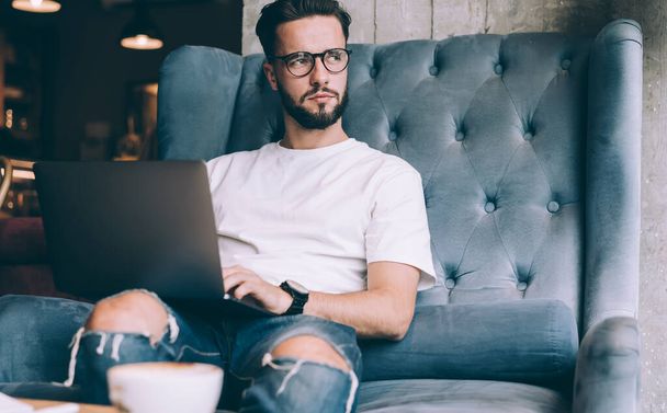 Pensive male IT software developer in optical spectacles for provide eyes correction thoughtful looking away during remote working on laptop technology, - 写真・画像
