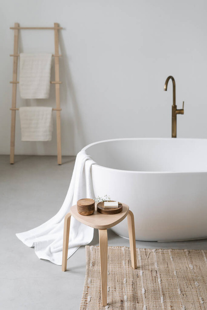 Wooden chair with soap bar and natural cosmetics for spa procedure close to tub. Vertical shot of white classic bathtub and home decor in modern interior. Comfortable bathroom in hotel - 写真・画像