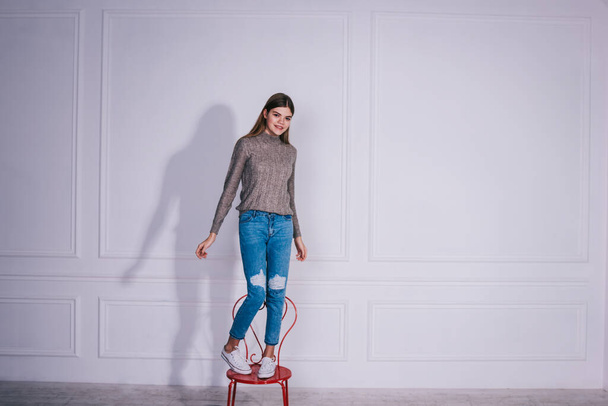 Full body of smiling female in casual outfit standing on metal chair in room with white wall and looking at camera with interest - Fotó, kép