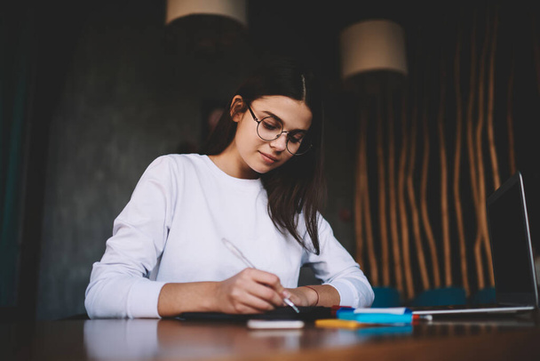 Thoughtful female freelancer in casual outfit and eyeglasses writing down thought in copybook while sitting at table with portable laptop and markers - Foto, afbeelding