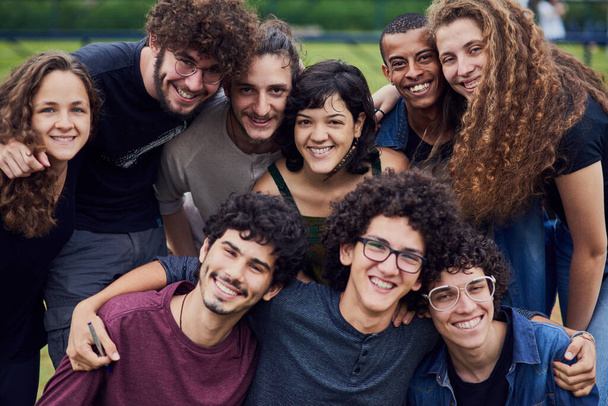 Retrato de um grupo de jovens estudantes de pé braços ao redor uns dos outros fora em um parque durante o dia. - Foto, Imagem