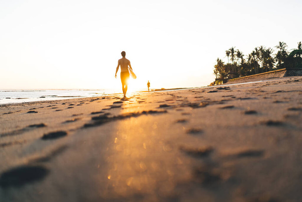 認識できない男性サーファーの地上レベルのバックビュー裸の胴はカラフルな太陽の下で夜にサーフボードを保持足跡と砂浜を歩く - 写真・画像